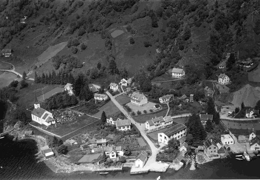 Bruvik 1955. Lengst til venstre løa på Kyrkje-Bruvik bnr. 10. Ovanfor den ligg løa på bnr. 2. Skulehuset midt på biletet ligg noko på Kyrkje-Bruvik og noko på Indre-Bruvik. Ovanfor skulehuset ser ein bedehuset. Til høgre for vegen opp frå kaien ligg eigedomen Solli med tekstilfabrikk (bnr. 23) og  Bruvik Handelslag (bnr. 24). Heilt til høgre nede ved sjøen ligg den gamle gjestgivarstaden og tingstova. Her var handel, kai og bakeri. Sjå bnr. 27.  Foto: Widerøe’s Flyveselskap A/S. 